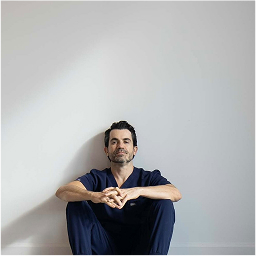 A man wearing navy blue scrubs sits on the floor against a plain white wall with his hands clasped and legs bent.