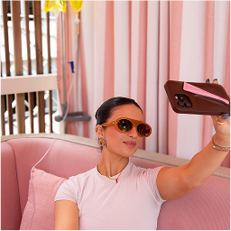 Woman wearing sunglasses takes a selfie with her phone while sitting on a pink couch in a room with pink and white curtains.