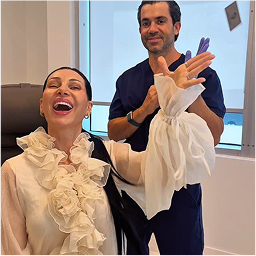 A woman in a ruffled blouse smiles and waves while seated; a man in scrubs stands behind her, putting on purple gloves.