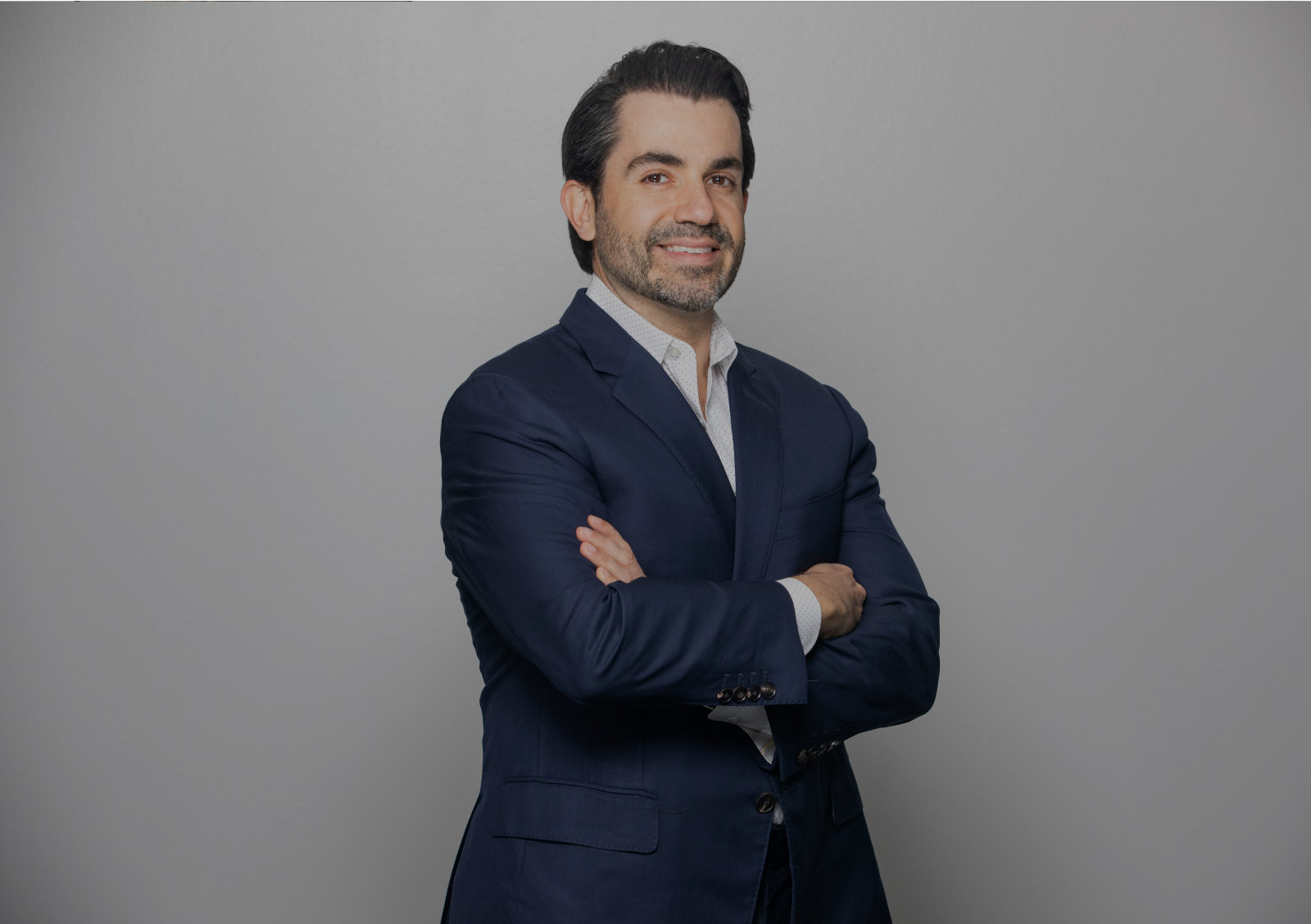Hombre con un traje azul oscuro con los brazos cruzados, de pie frente a un fondo gris claro liso.