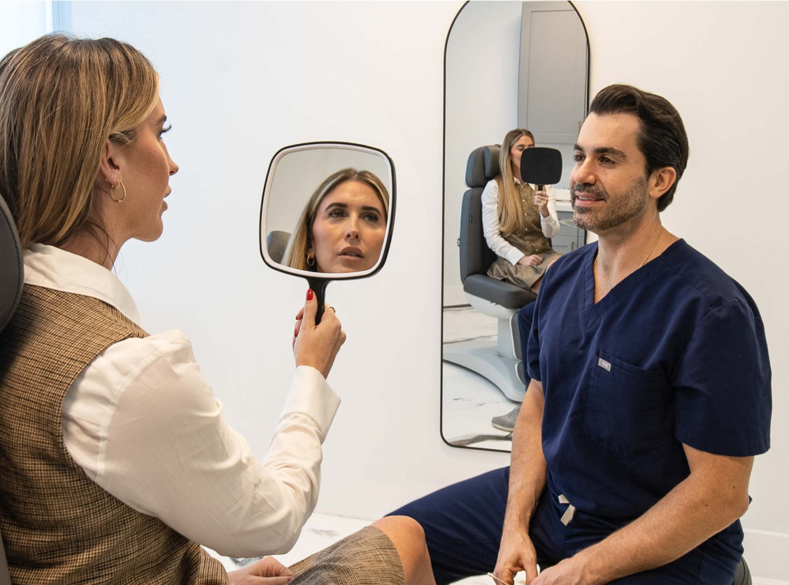 A woman holding a mirror looks at her reflection while speaking with a man in medical scrubs during a consultation in a modern clinic room.