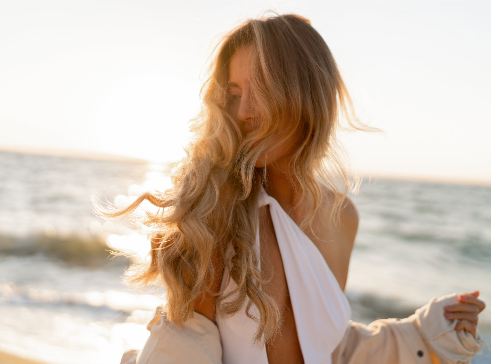 A woman with long wavy blonde hair stands on a beach at sunset, wearing a white halter top and holding a light jacket.