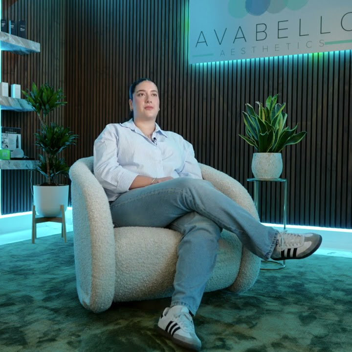 A person sits cross-legged in a modern, light-colored chair in an aesthetic clinic with plants and a glowing sign reading "AVABELLO AESTHETICS" in the background.