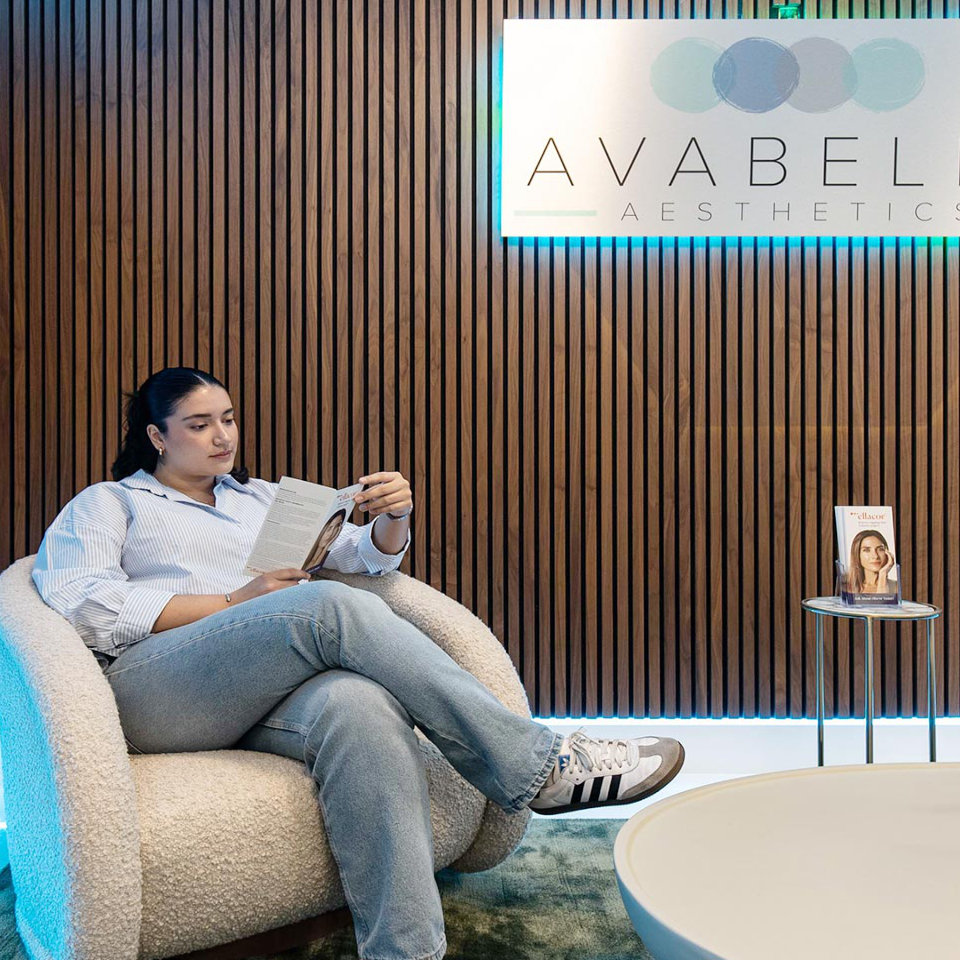 A person sits in a modern waiting area reading a brochure, with a sign reading "Avabel Aesthetics" and a small table holding a pamphlet nearby.