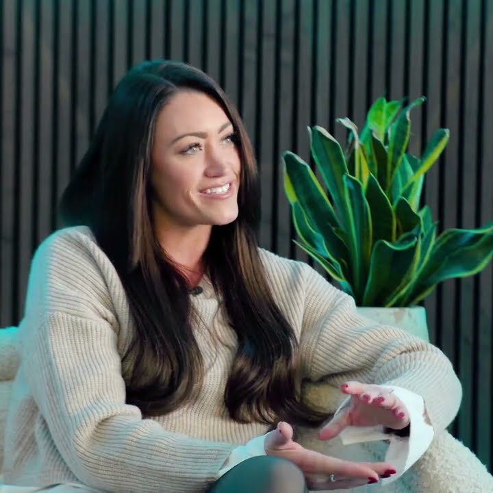 A woman with long dark hair sits on a chair, smiling and gesturing with her hands. A potted plant is visible in the background.