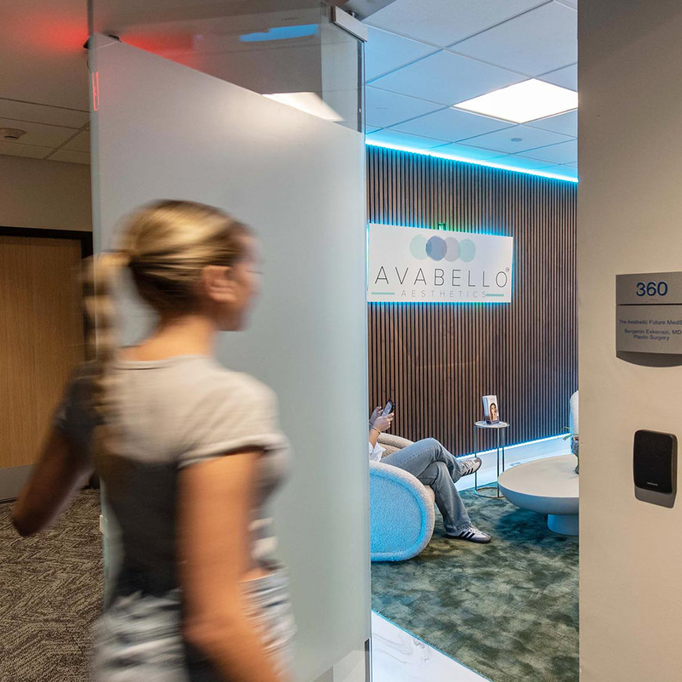 A person enters a modern aesthetics clinic waiting room, where two people are seated. The sign on the wall reads "Avabello Aesthetics.