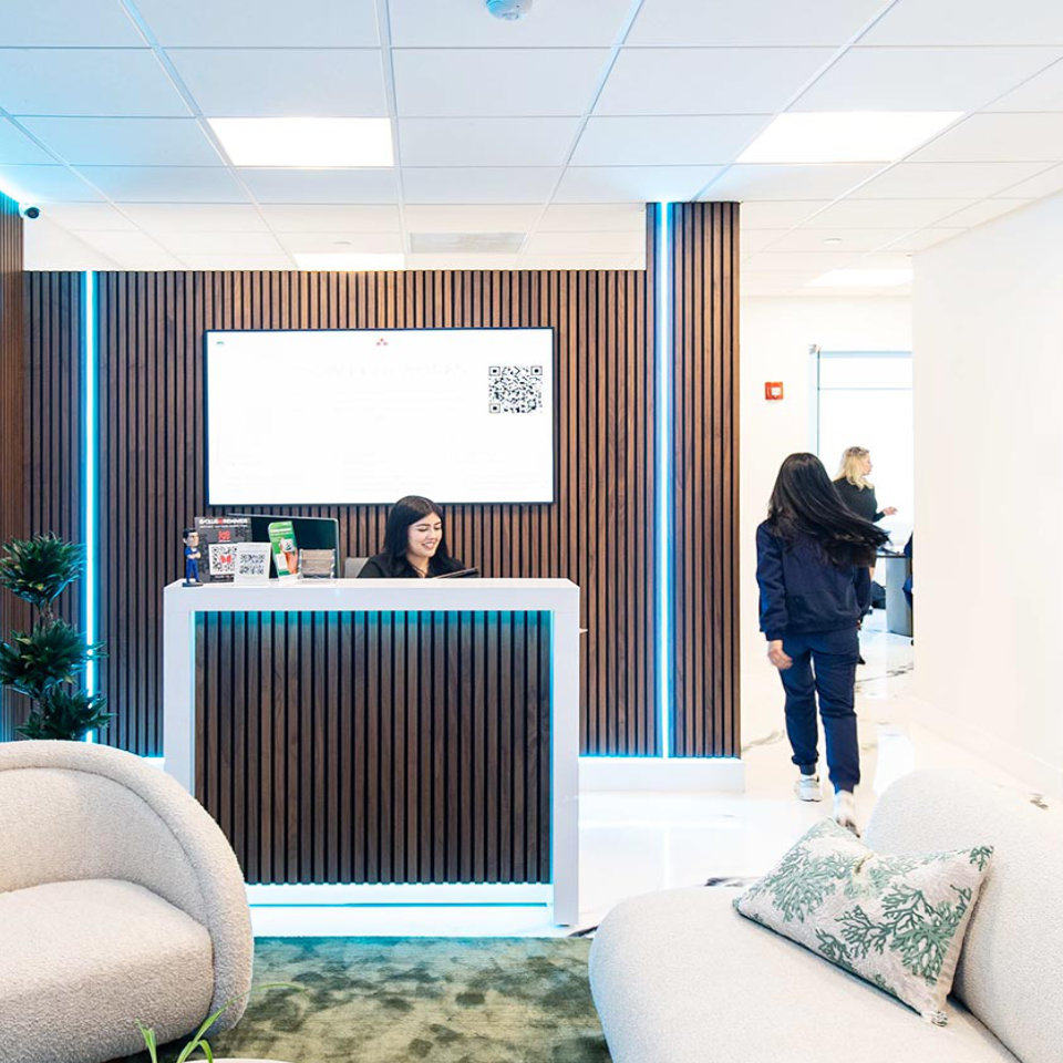A receptionist sits at a modern wooden front desk in a brightly lit office lobby, while a woman walks toward a hallway.