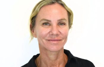 A middle-aged woman with light hair and blue eyes smiles slightly, wearing a black shirt, against a plain white background.