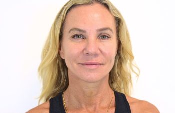 A middle-aged woman with blonde hair, light eyes, and a neutral expression, wearing a sleeveless black top and a gold necklace, standing against a plain white background.