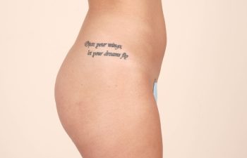Side view of a person's torso with a tattoo on the ribcage reading "Open your wings, let your dreams fly" against a plain background.