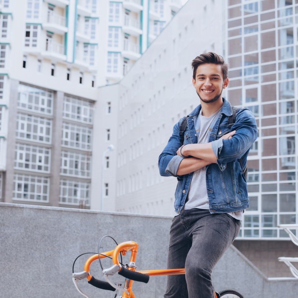 Man riding a bicycle outside. Smiling handsome young guy with beard in jacket looking at camera. He is standing on a bike with crossed hands. Urban background