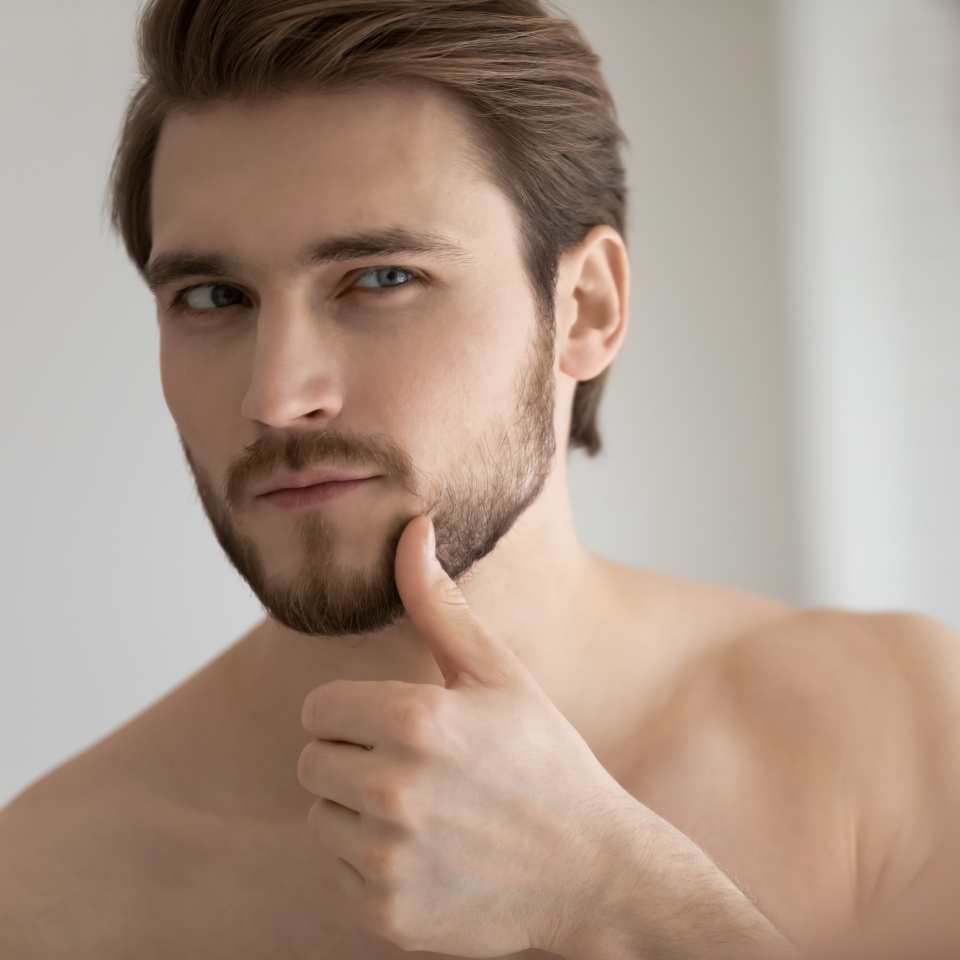 Close up headshot mirror reflection satisfied handsome young man touching beard, standing in bathroom, applying aftershave lotion, enjoying skincare procedure, morning routine concept