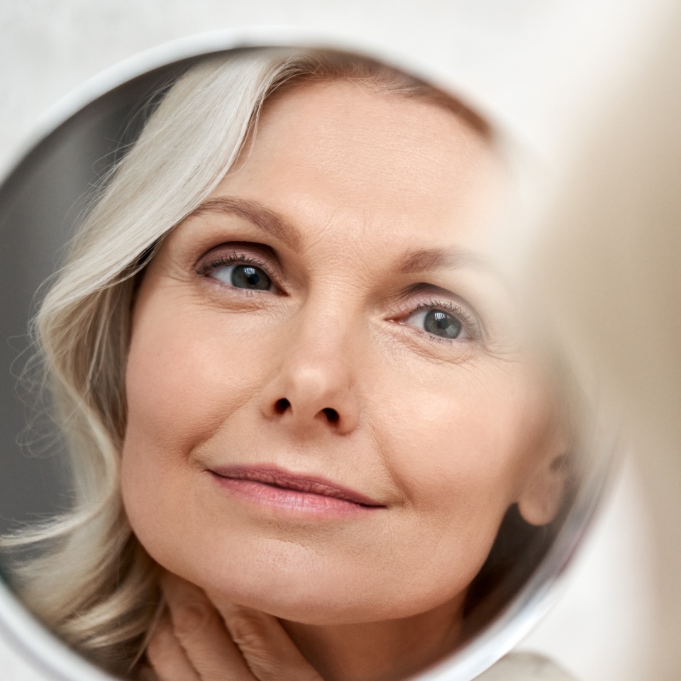 Happy 50s middle aged woman model touching face skin looking in mirror reflection. Smiling mature old lady pampering, healthy moisturized skin care, aging beauty, skincare treatment cosmetics concept.