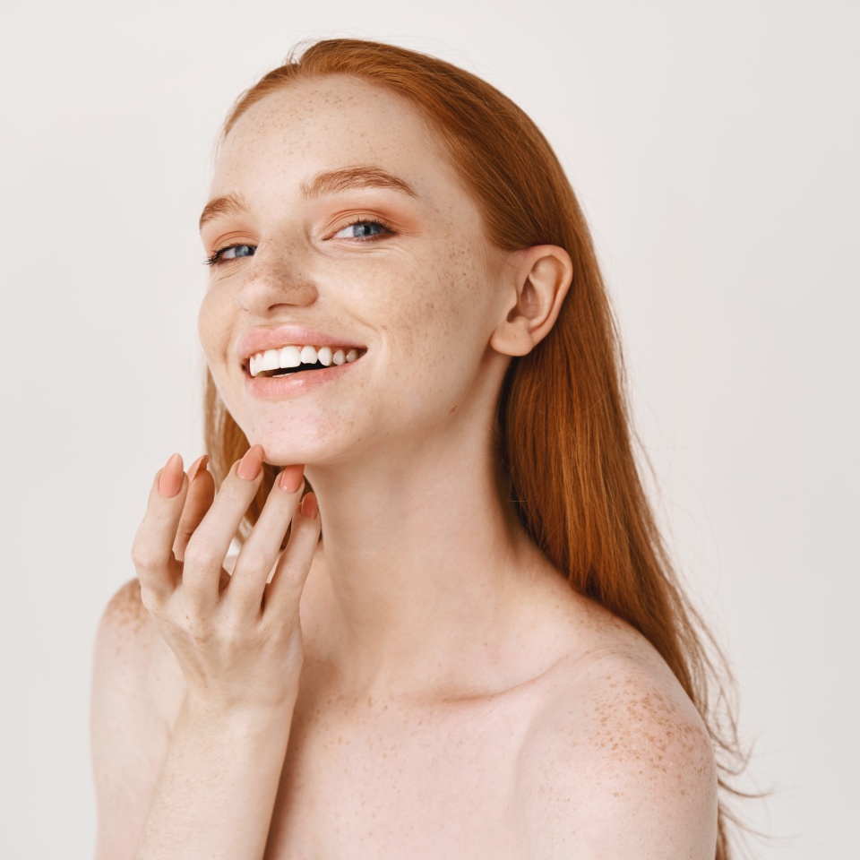 Close-up of smiling redhead woman with pale skin and freckles touching soft, perfect face, using skincare cream, standing over white background.