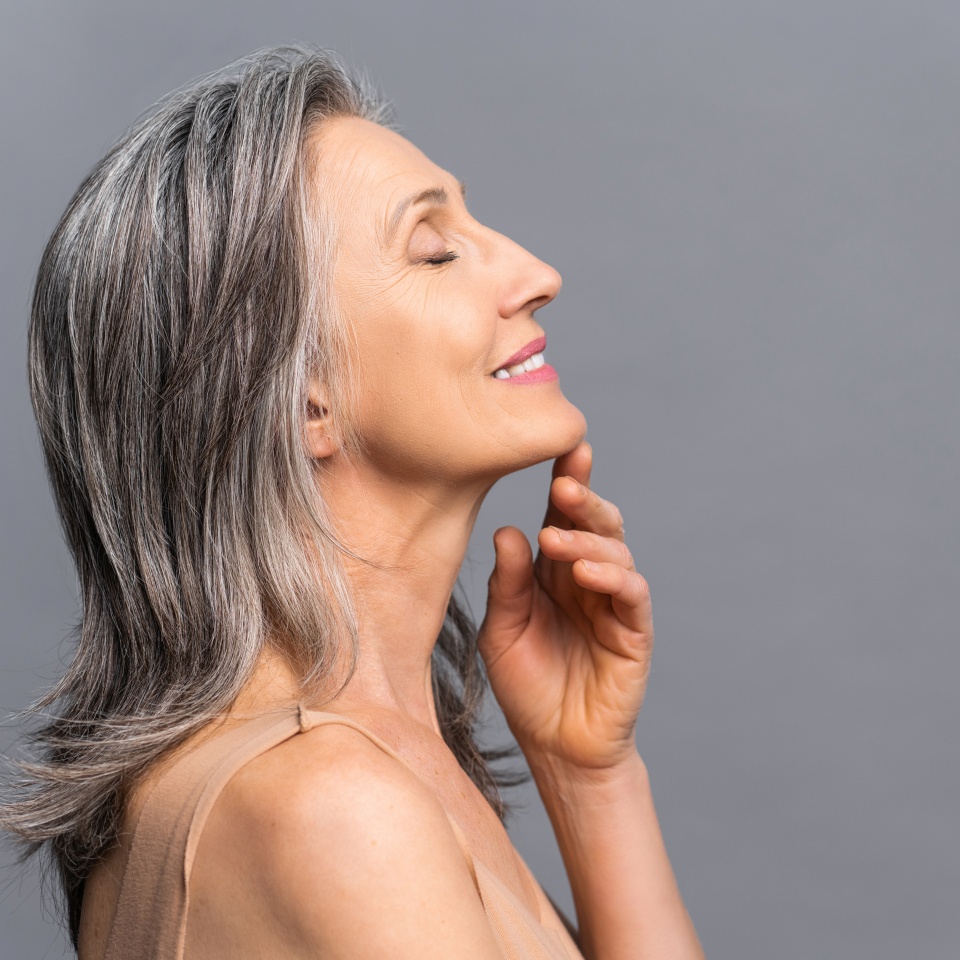Sensual calm elderly woman stands in profile isolated on gray, smiling with eyes closed, touching chin with hand, gray-haired charming lady enjoying smooth of fresh skin after spa procedure