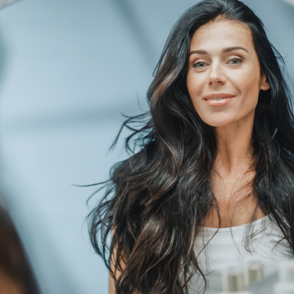 Beautiful Caucasian Middle Aged Woman Looks into Bathroom Mirror, Enjoys Her Looks. Concept for Happiness, Wellbeing, Natural Beauty and Organic Skin Care Products