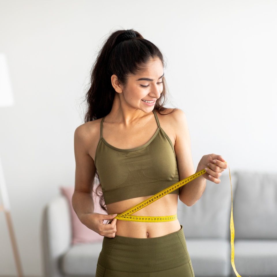 Concepto de pérdida de peso exitosa. Joven india en ropa deportiva midiendo la cintura con cinta métrica en casa. Señora millennial sintiéndose satisfecha con el programa de adelgazamiento en interiores  