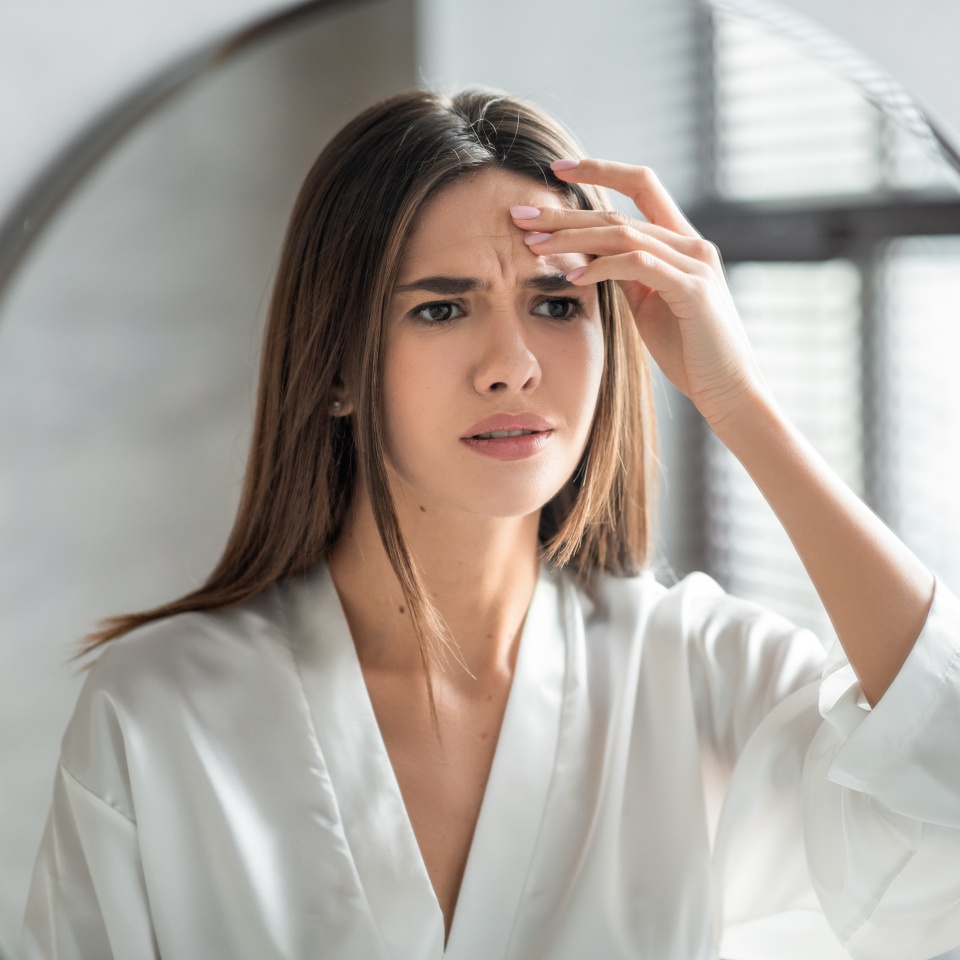 Unhappy Young Woman Looking In Mirror And Touching Wrinkles On Her Face, Attractive Millennial Female Standing In Bathroom And Examining Skin Fine Lines, Selective Focus On Reflection, Closeup