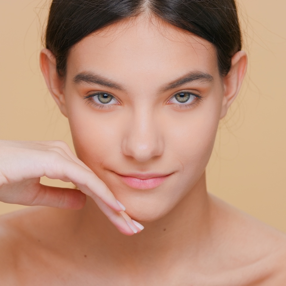 A young person with straight dark hair gazes at the camera, resting their chin on their hand against a plain beige background.