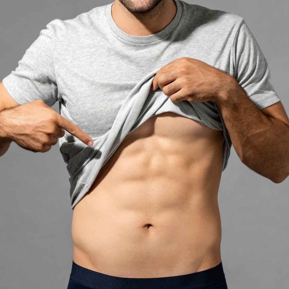 Muscular male model lifting up t-shirt to show abs while standing on gray isolated background in studio