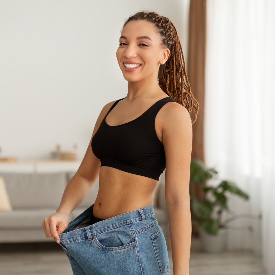 Éxito en la pérdida de peso. Señora negra delgada y feliz que usa jeans viejos de gran tamaño que muestran el resultado de la dieta de pie sonriendo a la cámara en casa. Concepto de motivación para adelgazar con éxito  