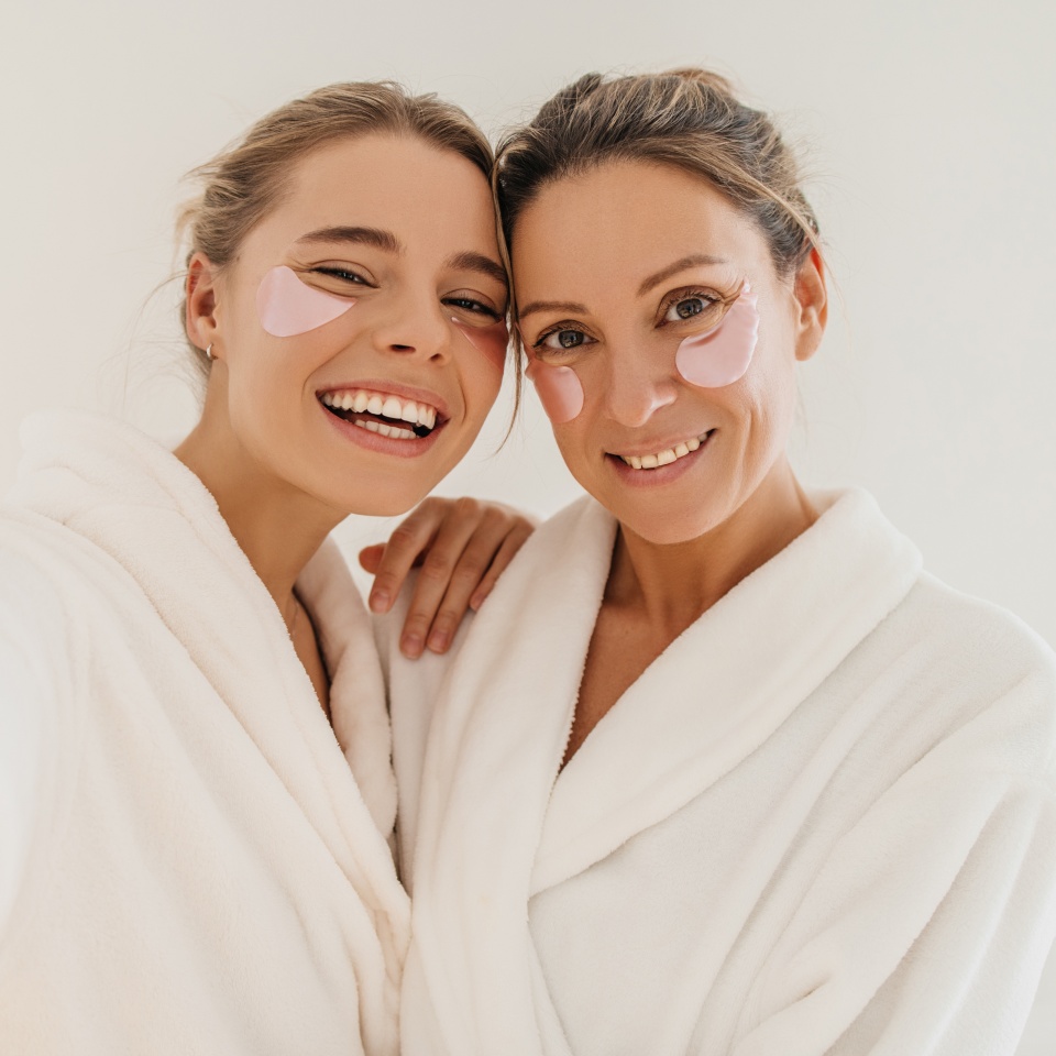 Happy caucasian mother with daughter wearing hydrogel patches smiling looking at camera on white background. Blondes wear robes to take care of their skin. Cosmetic natural trend.