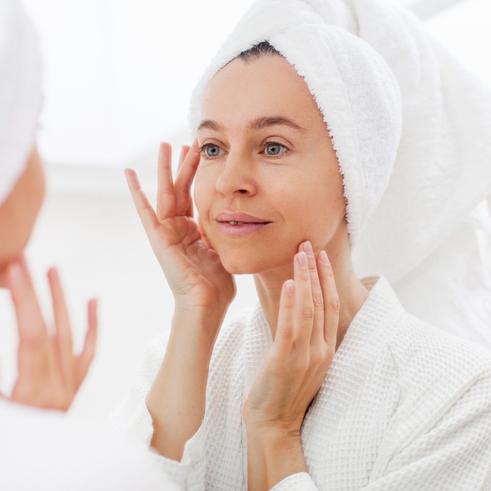 Gorgeous mid age adult 40 years old blonde woman standing in bathroom after shower touching face, looking at reflection in mirror doing morning beauty routine. Older dry skin care concept