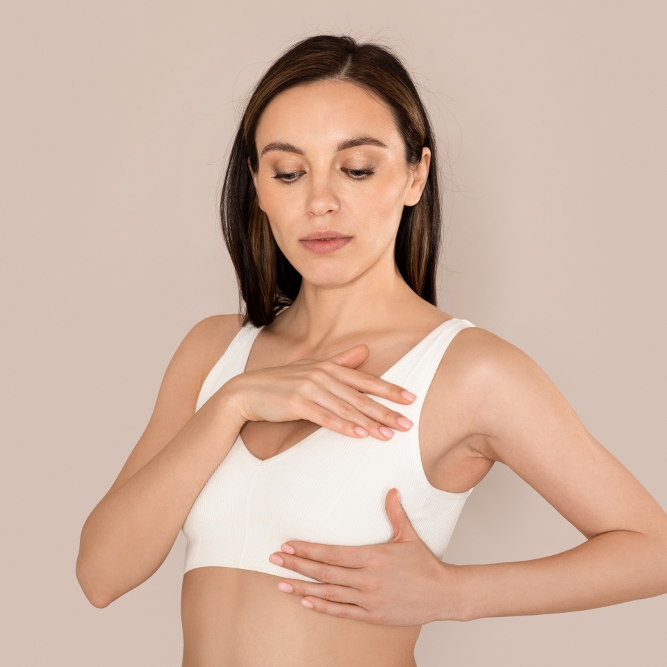 Breast Self-Exam, BSE. Beautiful young woman wearing underwear white top checking her breast on beige studio background. Concerned lady make screening, cancer prevention concept