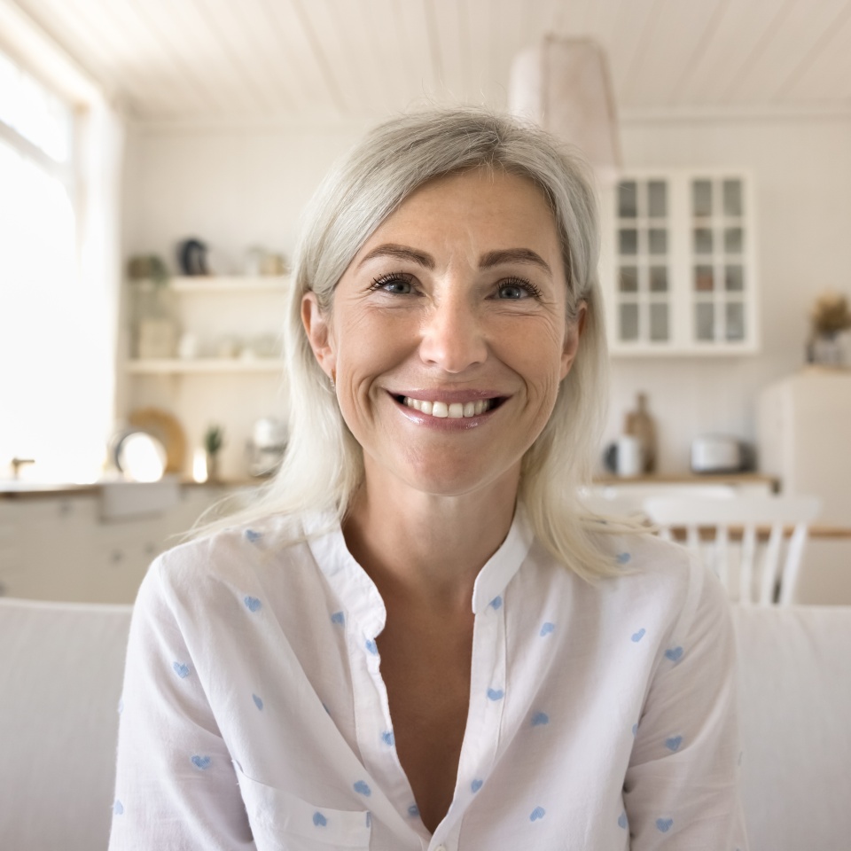 Positive pretty blonde mature woman home head shot portrait. Happy senior lady talking on online video conference call, looking at camera with toothy smile, sitting on couch with kitchen in background