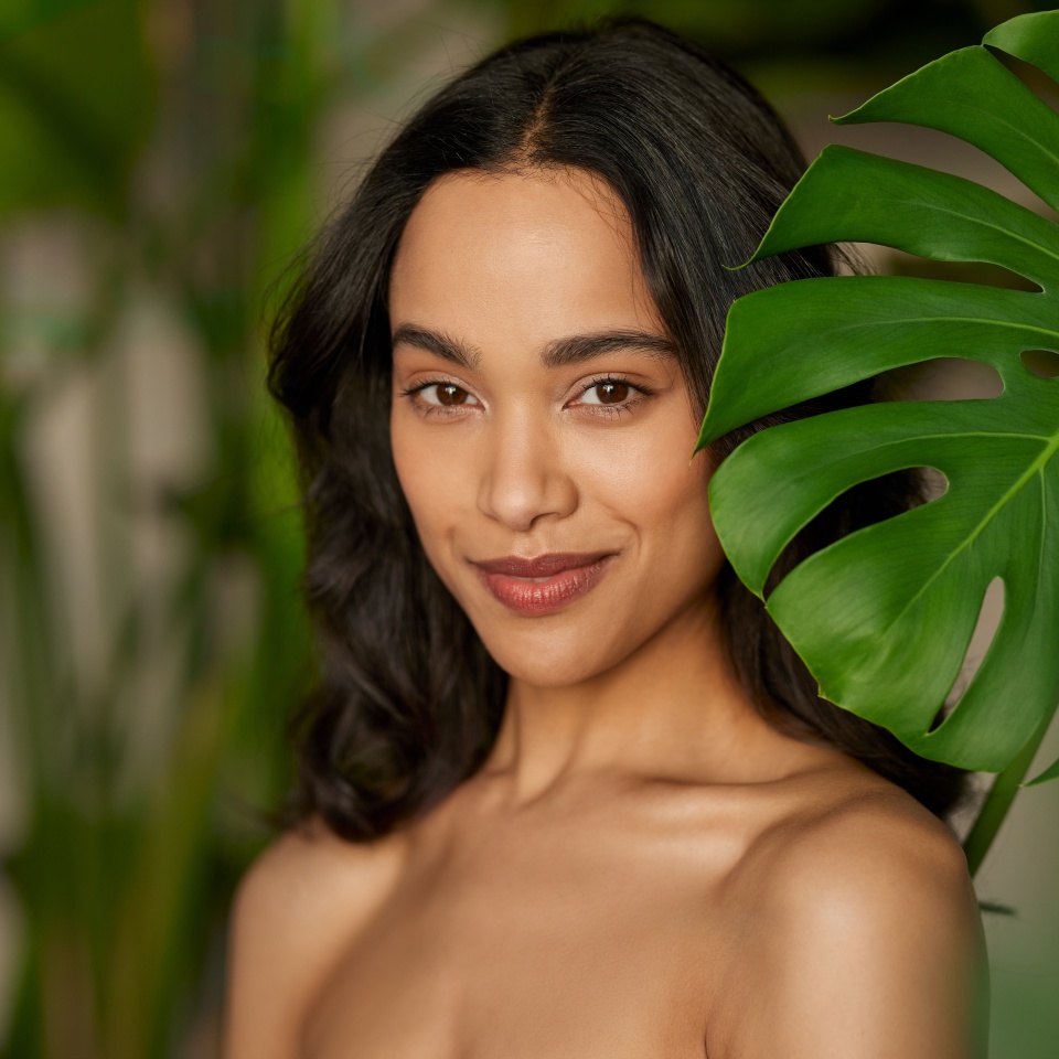 Portrait of beauty young woman smiling near green monstera leaf while looking at camera. Hispanic girl with natural makeup and perfect skin standing behind big green leaves with copy space.