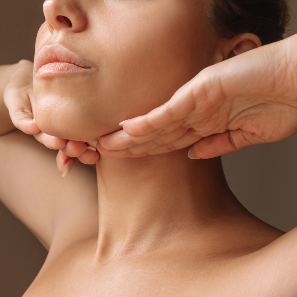 Toma recortada de una joven caucásica tocándose debajo del mentón con las manos masajeando su rostro sobre un fondo marrón oscuro. Rejuvenecimiento, lifting facial, facefitness. Ejercicios desde el segundo mentón, cuello de pelícano  