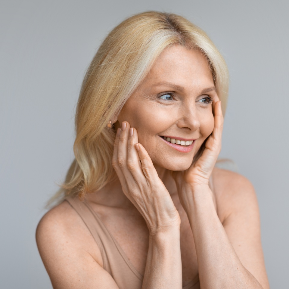Beautiful mature woman looking aside at copy space and touching cheeks, smiling attractive aged lady enjoying skincare treatments, standing over grey studio background, panorama