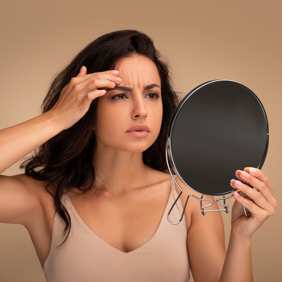 Fine lines and wrinkles, premature skin aging concept. Concerned upset young eastern woman looking in mirror, touching her forehead, checking aging signs on her face, beige background