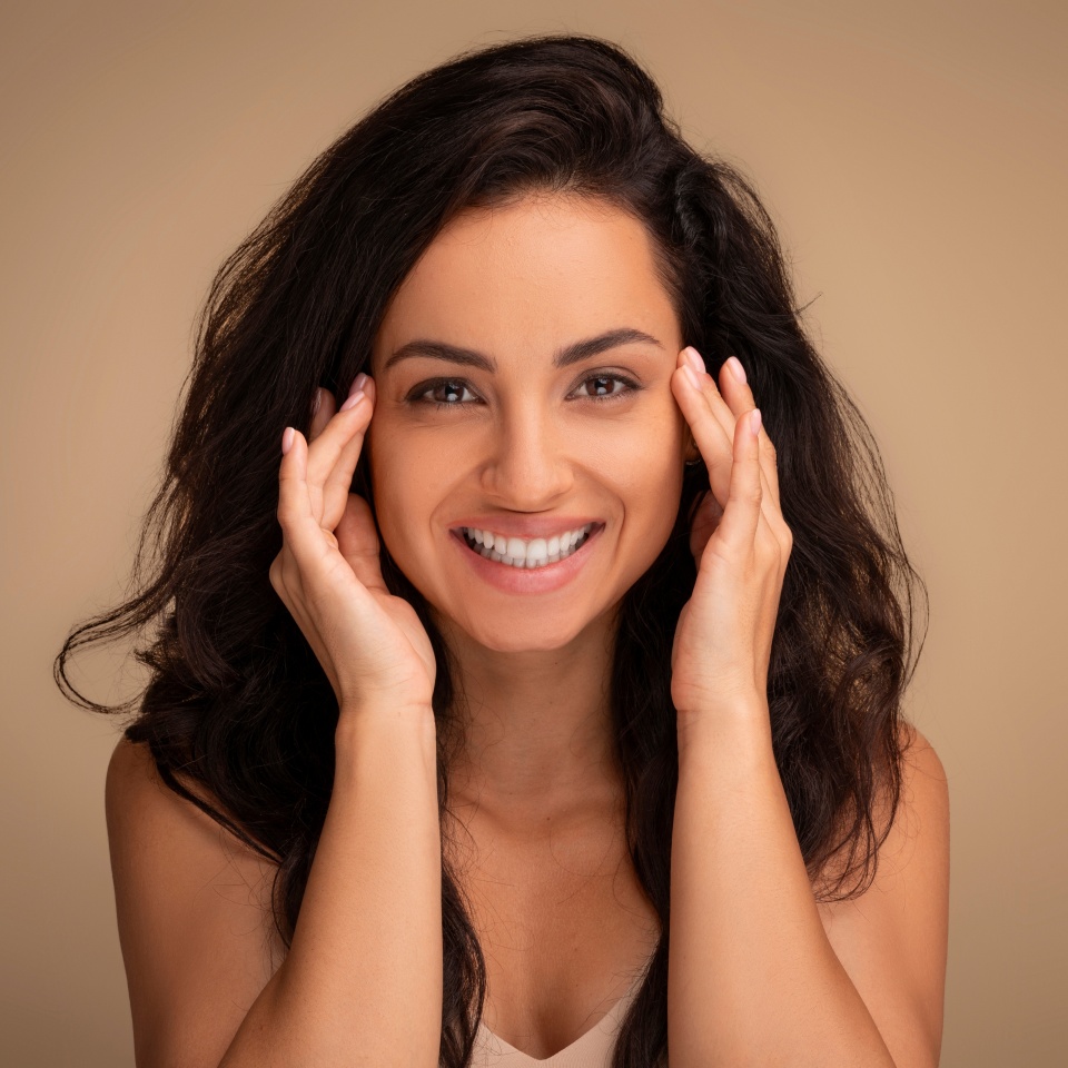 Anti-aging face care treatment, aesthetic procedures concept. Smiling attractive brunette long-haired young woman touching face skin temples, closeup, beige studio background,, copy space