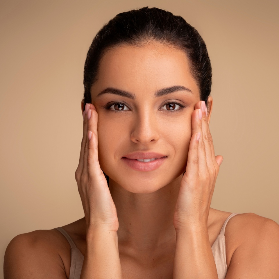 Antiage skincare. Beautiful millennial woman touching skin near eyes and looking at camera, lady with flawless skin standing over beige studio background, closeup portrait, panorama, copy space