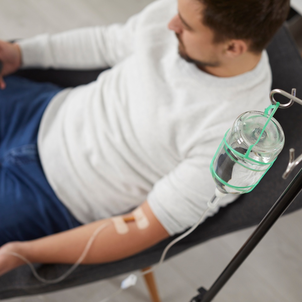 Top view photo of a man sitting in the chair and using the smartphone while receiving IV drip infusion and vitamin therapy in his blood. Person receiving injection therapy. Healthcare concept.
