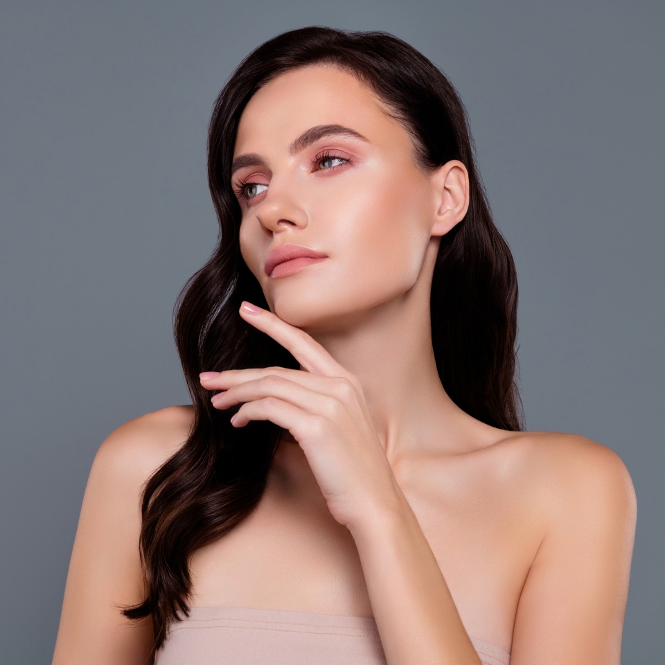Woman with long dark hair poses against a grey background, looking to the side with her hand resting near her chin, wearing a strapless top.