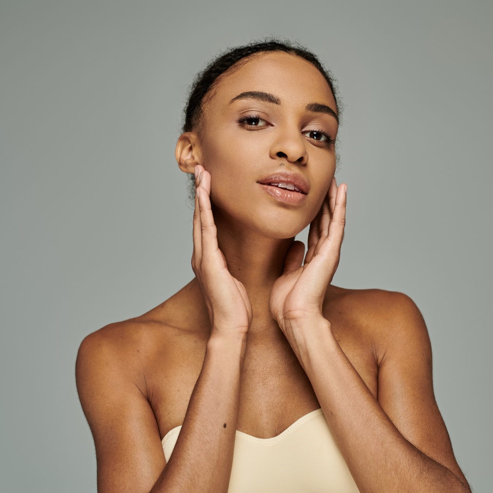 A beautiful young African American woman in a strapless top, gently touching her face with both hands, on a grey background.