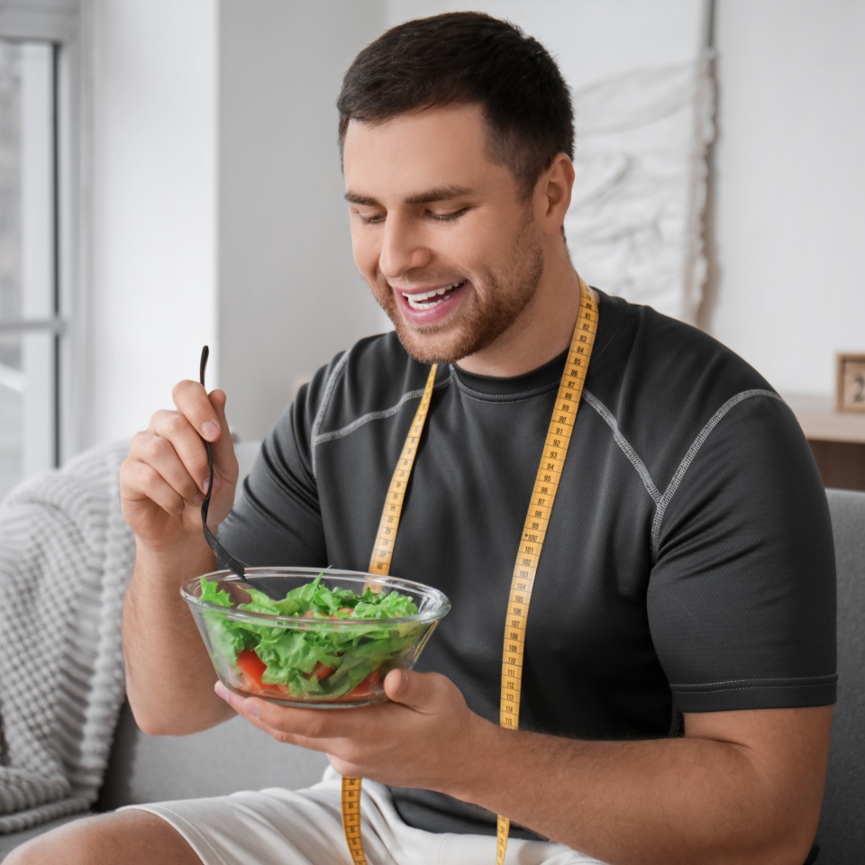 Un hombre con una cinta métrica alrededor del cuello está sentado en el interior, sonriendo mientras come una ensalada de un cuenco de cristal con un tenedor.