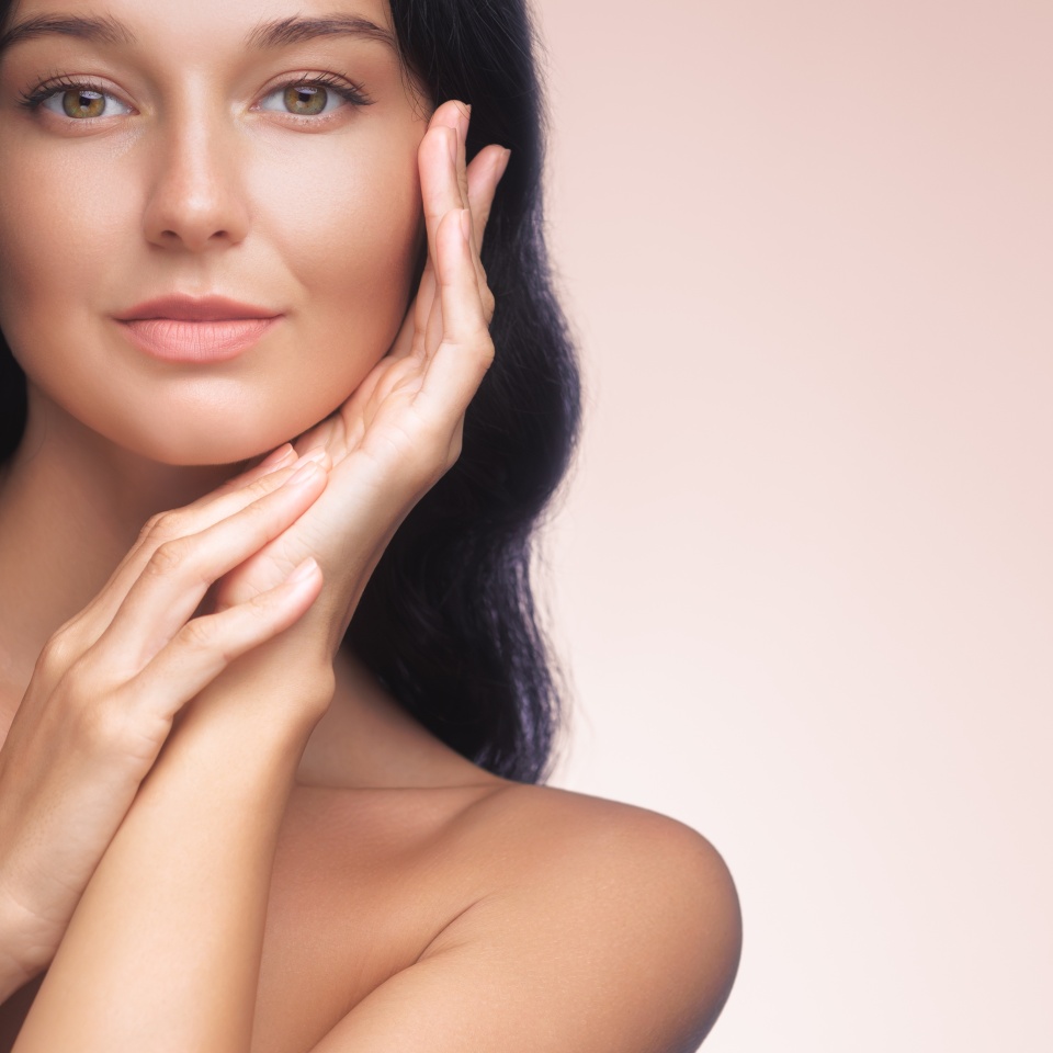 An elegant Asian woman with long black hair gently touches her face, showcasing her flawless skin against a soft pink background. Ideal for beauty, skincare, and cosmetic advertisements, emphasizing