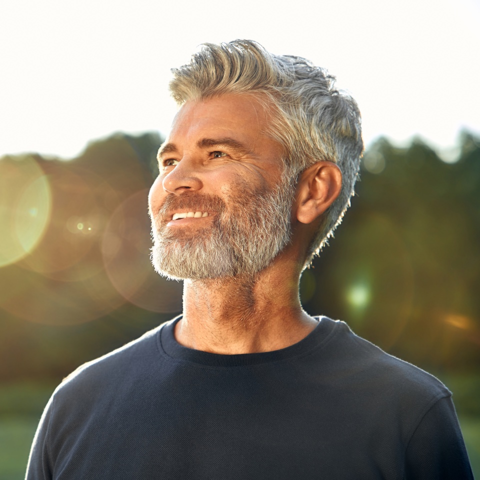 A smiling middleaged man, characterized by his gray hair and beard, stands outdoors, illuminated by the warm light of the sunset, radiating positivity, contentment, and a deep sense of tranquility