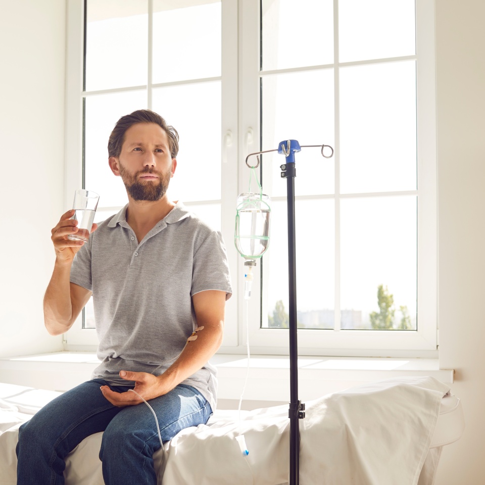 Drip therapy. Sick, dehydrated or hungover man receives IV vitamin infusion and drinks water in hospital. Millennial Caucasian male patient is connected to drip and sits on couch in hospital ward.