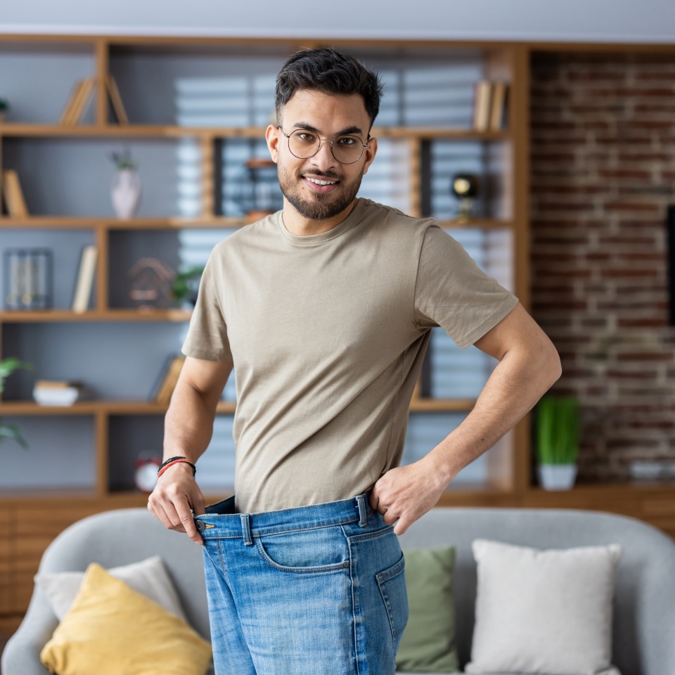 Retrato de un joven indio sonriente de pie en casa frente a la cámara con vaqueros y mostrando el resultado de la dieta, el ejercicio y la pérdida de peso.