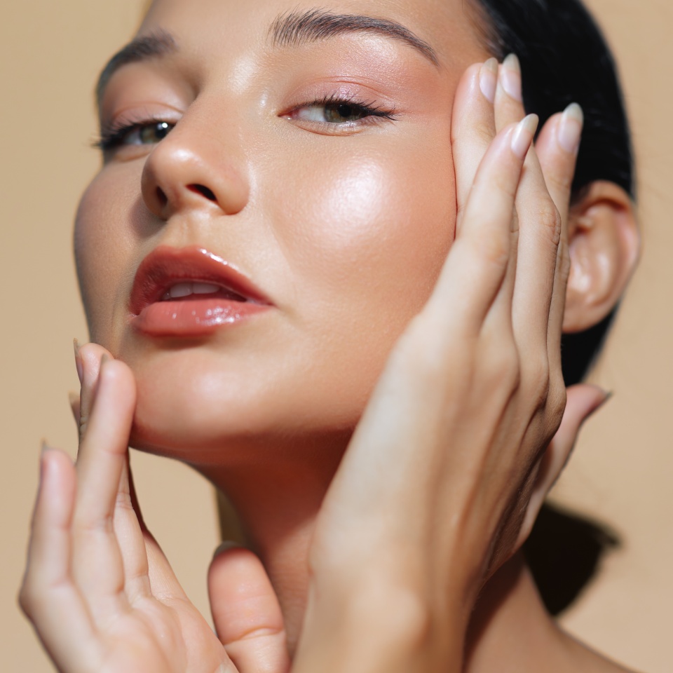 Impresionante primer plano de una mujer latina con piel radiante y maquillaje natural, acariciando con gracia su rostro con ambas manos sobre un cálido fondo beige. Ideal para anuncios de cuidado de la piel, editoriales de belleza y