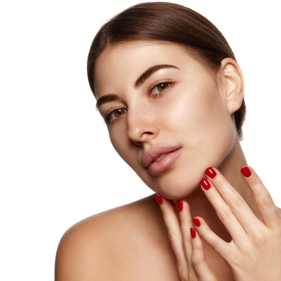 A woman with natural makeup and red nail polish touches her face and chin, posing against a white background.
