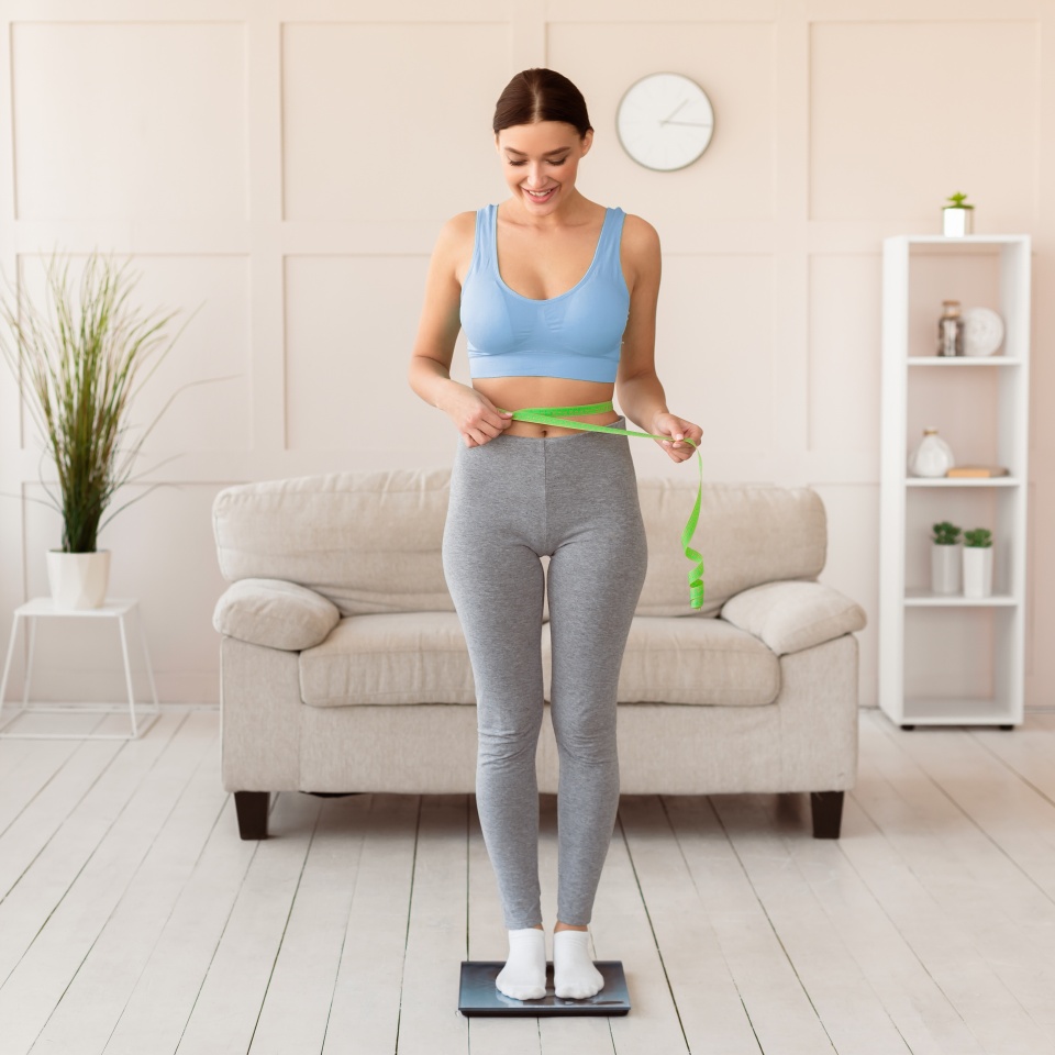 Una mujer en ropa deportiva está de pie sobre una báscula, sonriendo y midiéndose la cintura con una cinta métrica en una sala de estar.