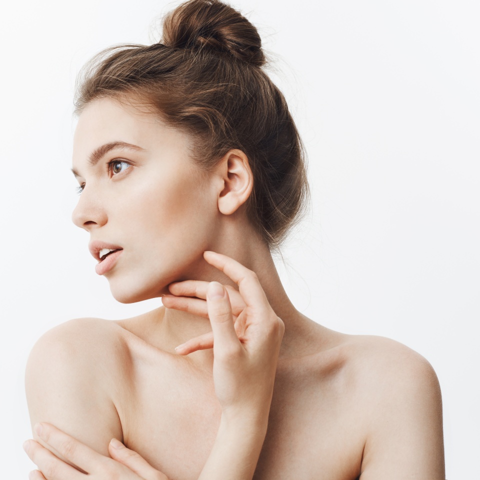 Close up portrait of young female brunette student girl with bun hairstyle and baked shoulders looking aside with calm face expression touching jawline with fingers. Beauty and health