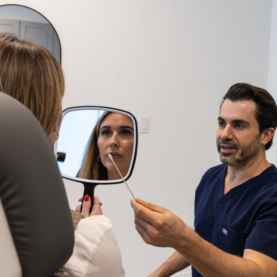 A doctor in navy scrubs holds a swab while a woman looks at her reflection in a handheld mirror during a consultation in a medical office.