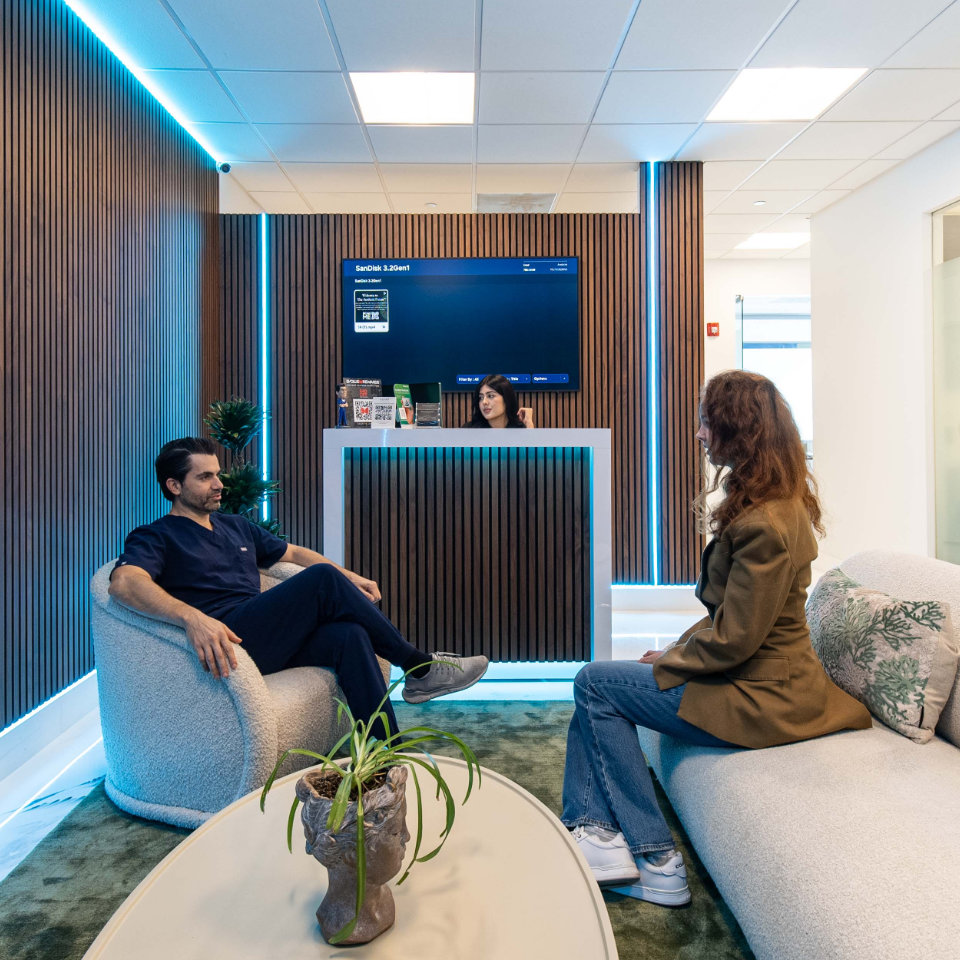 A man and a woman sit in a modern waiting area with wood paneling, while a receptionist stands behind a counter with a digital screen above her.