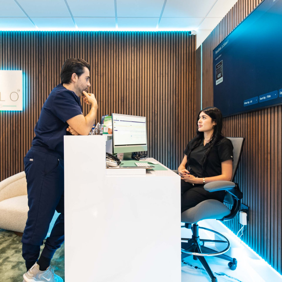 A man in scrubs stands by a desk talking to a seated woman in an office with wood panel walls, a computer, and a large screen on the wall.